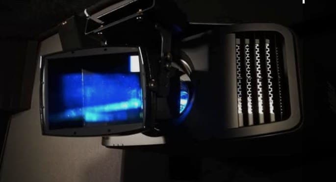 A high-end home cinema projector with an anamorphic lens attachment, glowing faintly in a darkened media room
